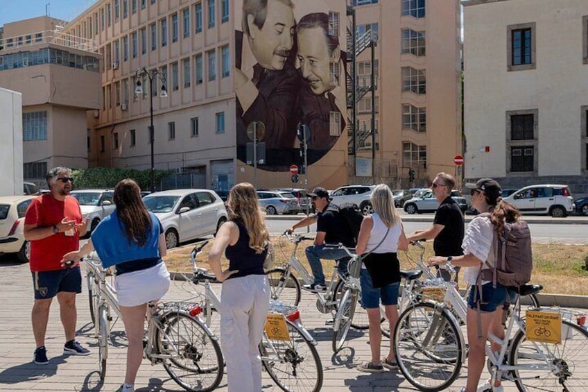 Un grupo de personas en bicicleta en un entorno urbano con un gran mural de fondo. 1