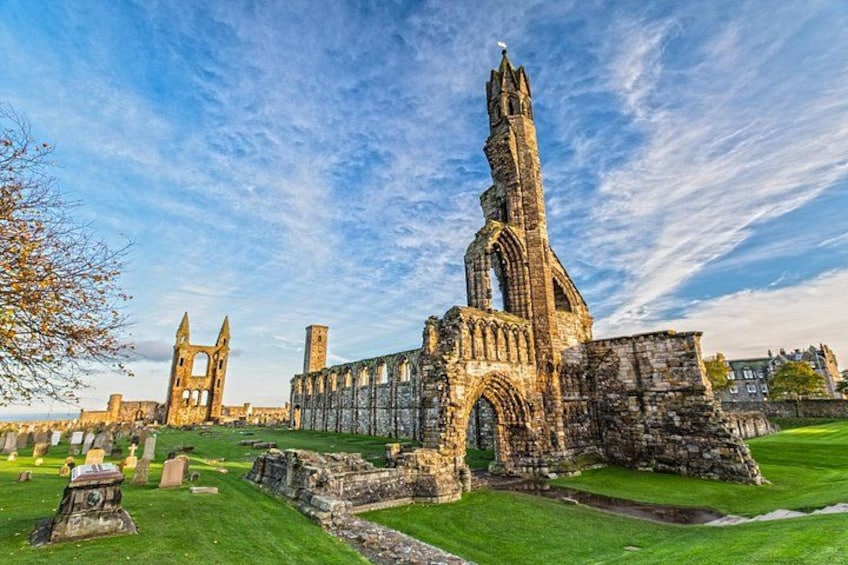 A historic stone structure with a tall spire and arches, surrounded by a grassy area with other ruins. 1