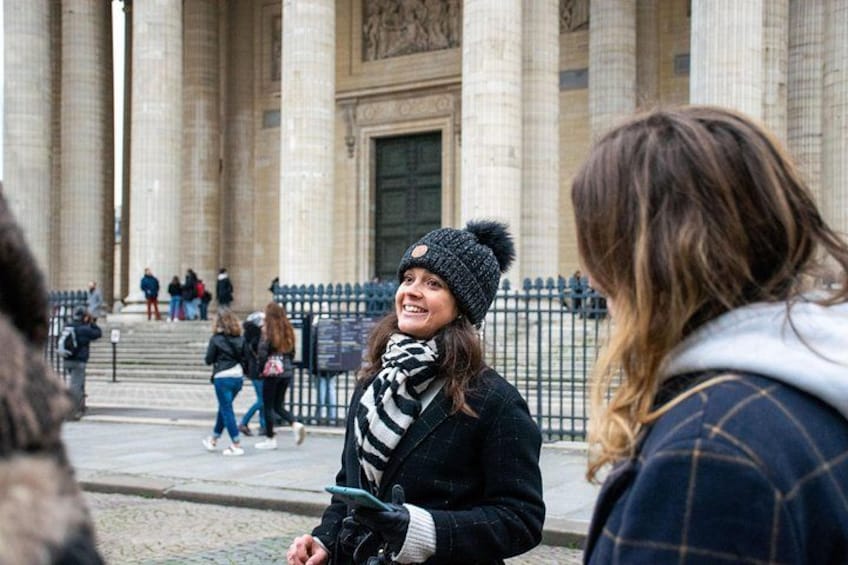 Una persona con una giacca nera e una sciarpa a righe, in piedi di fronte a un grande edificio con colonne. 3