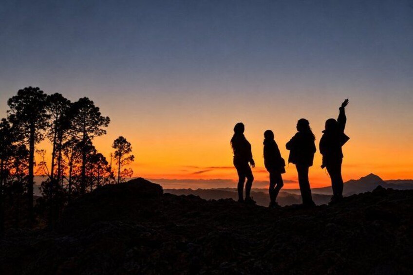 Solnedgang med fire silhuetter af mennesker stående på et klippefyldt område med træer. 1