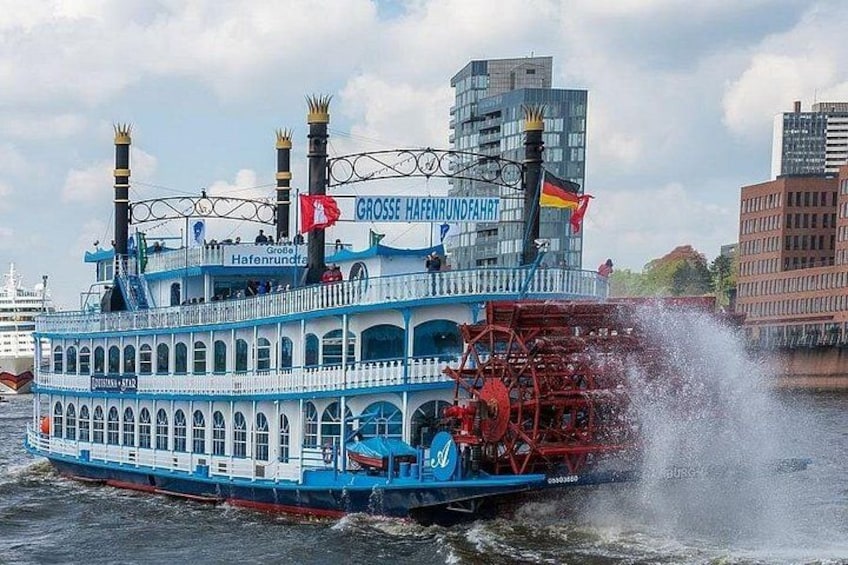 Een blauwe rivierboot met een rood peddelrad, genaamd 'Grosse Hafenfahrt', ligt aangemeerd aan een steiger, met gebouwen op de achtergrond. 3