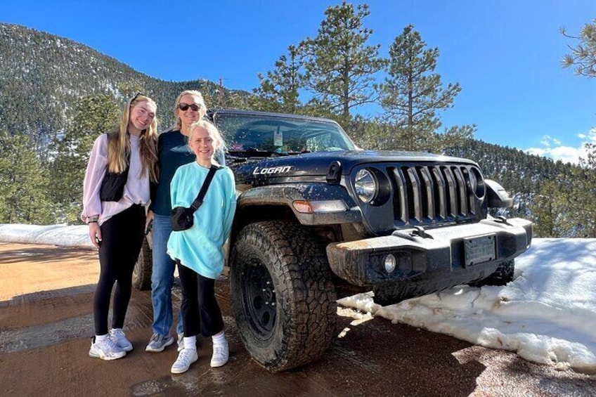 A Jeep Wrangler with a large off-road tire, a front grille, and a license plate, parked on a snowy path with a mountainous background. 4