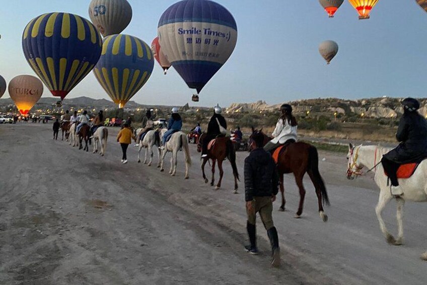 A group of people riding horses along a dirt road with hot air balloons in the background. 5