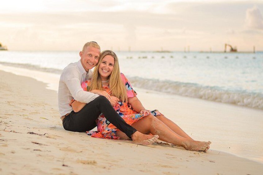 A couple sitting on a beach with the ocean and a pier in the background. 1