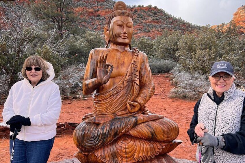 Une grande statue en bois représentant une figure assise, les mains jointes dans une posture méditative, entourée de formations rocheuses rouges et d’une végétation clairsemée. 2