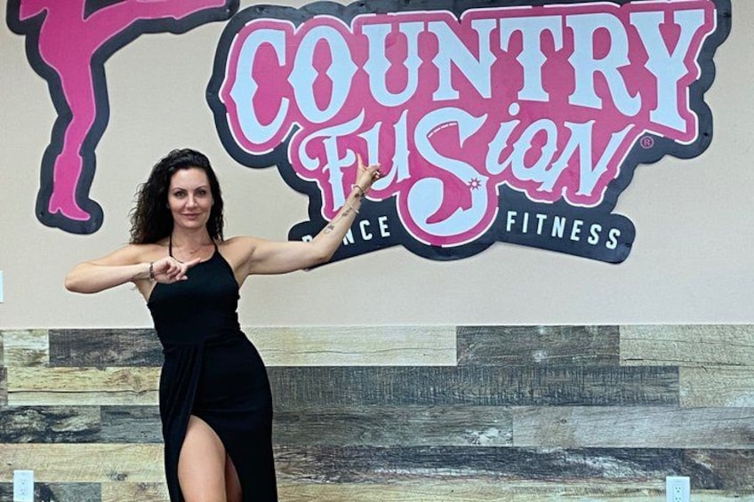 En person i en svart klänning poserar framför en skylt med texten ”Country Fusion Fitness”. 2