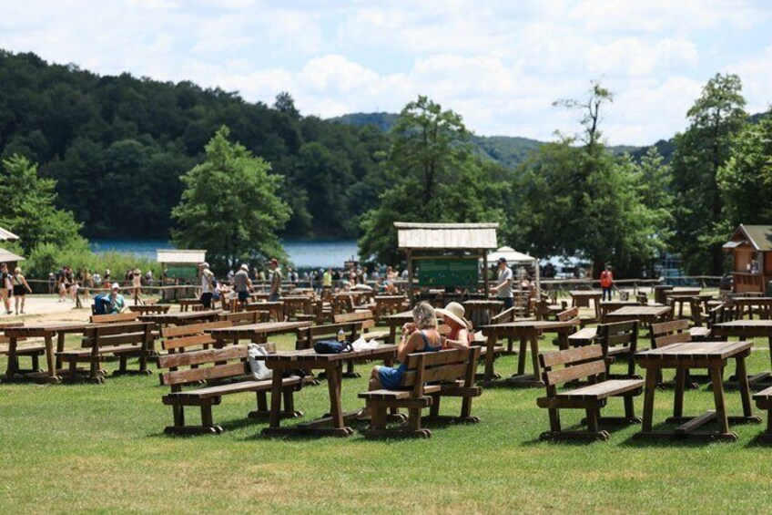 Picnic tables and people in a park with a lake and trees in the background. 3