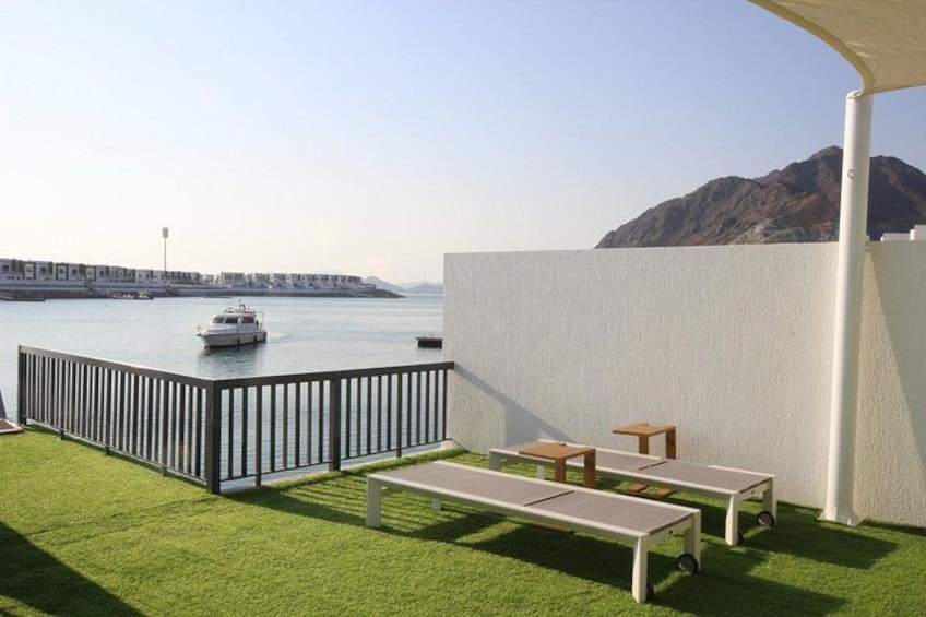 A balcony with artificial grass, two sun loungers, a small table, and a view of a body of water with a boat and distant buildings. 2