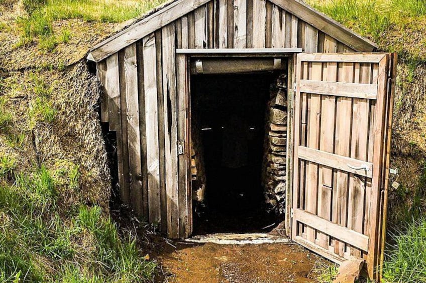A rustic wooden shed with a partially open door, surrounded by grass and a stone wall. 2