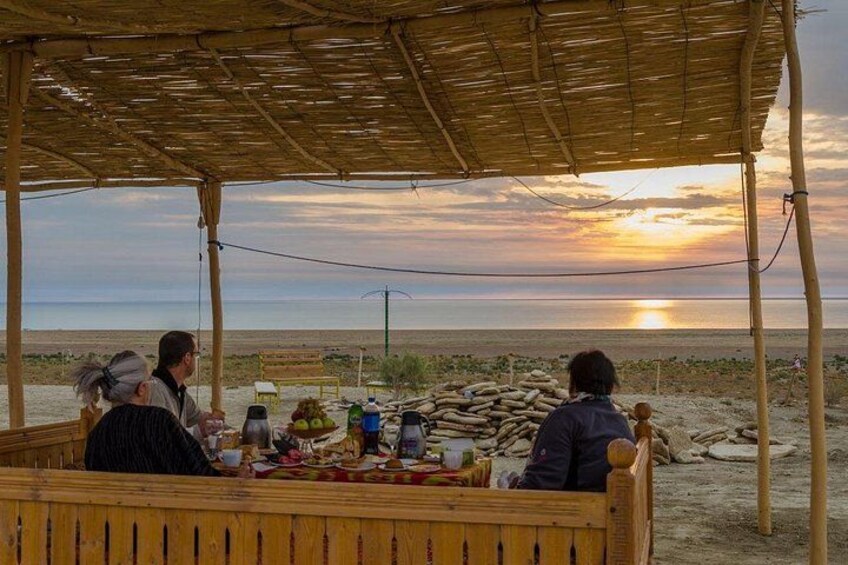 A wooden bench with people sitting, a table with food and drinks, and a sunset over a beach. 2