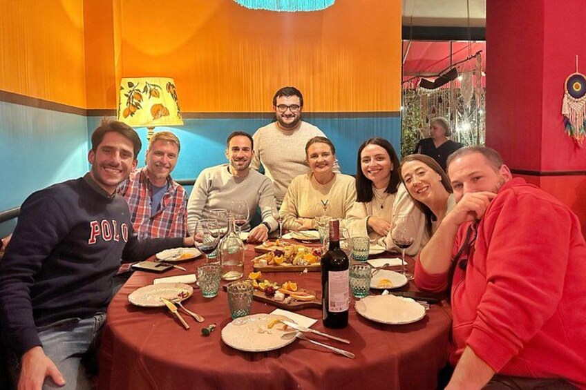 Un grupo de personas sentadas alrededor de una mesa con comida y bebidas, en una habitación con paredes coloridas y una lámpara. 1