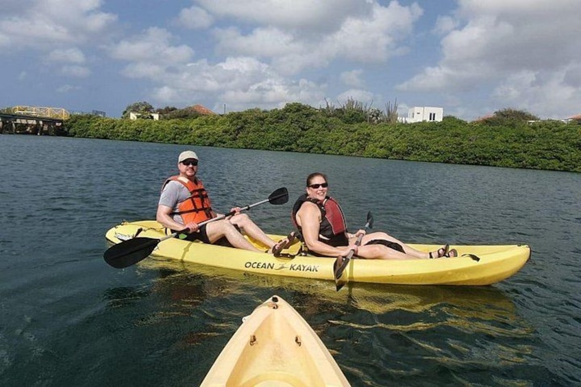 Two people kayaking on a calm river, surrounded by greenery and a bridge in the background. 3