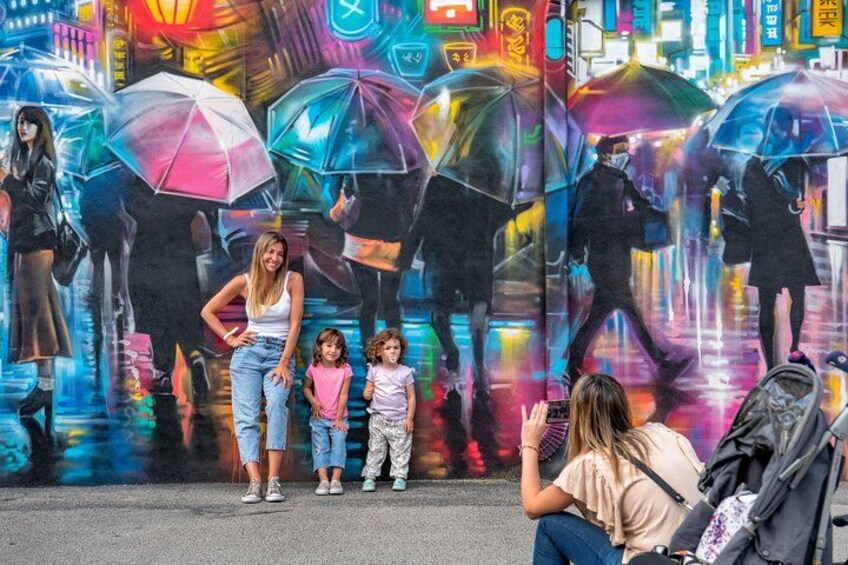 Una donna con due bambini davanti a un murale colorato con ombrelli. 5