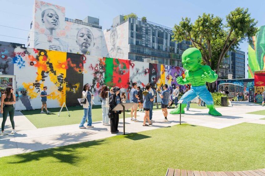 Uno spazio pubblico con una grande scultura verde, un murale con motivi colorati e una zona erbosa. 1