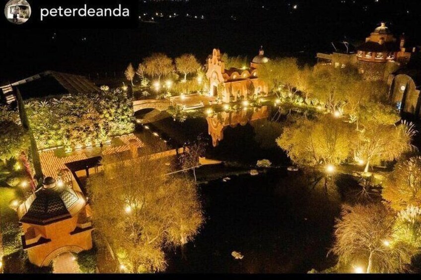 A nighttime view of a well-lit garden with pathways, a central fountain, and surrounding buildings. 5