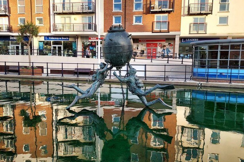 Eine Skulptur einer Krabbe in einem Brunnen, mit Spiegelungen von Gebäuden und einem Fußweg im Hintergrund. 3