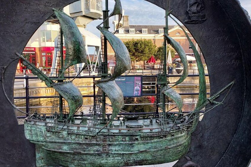 Eine Schiffs-Skulptur mit Segeln, einem grünlichen Patina-Überzug und einer Stadtansicht im Hintergrund. 4