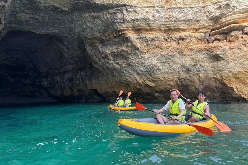 Två personer paddlar kajak i klart, turkosfärgat vatten med en klippig grotta i bakgrunden. 2
