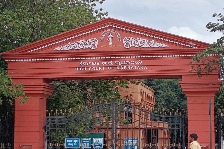 La entrada de la Alta Corte de Karnataka, con un arcojón rojo con inscripciones y una entrada protegida por una verja. 3