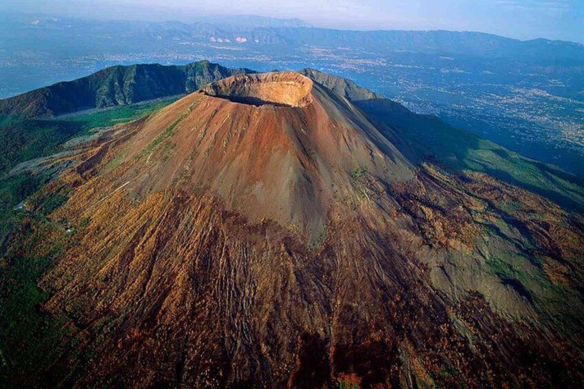 A volcanic mountain with a crater, surrounded by green vegetation and distant cityscape. 1