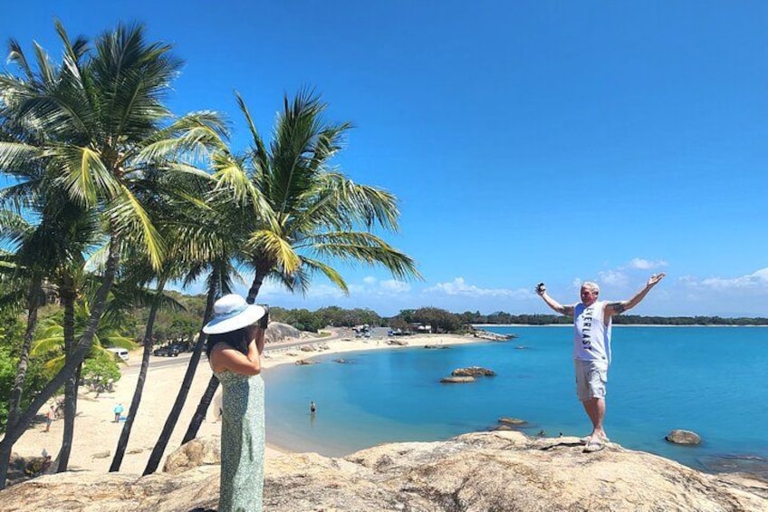 En strand med palmer, klart blåt vand og en person stående på en klippe. 2