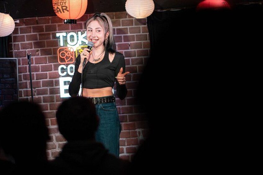A person speaking into a microphone on a stage with a brick wall background and a sign that reads 'TOKYO COMEDY CLUB'. 3