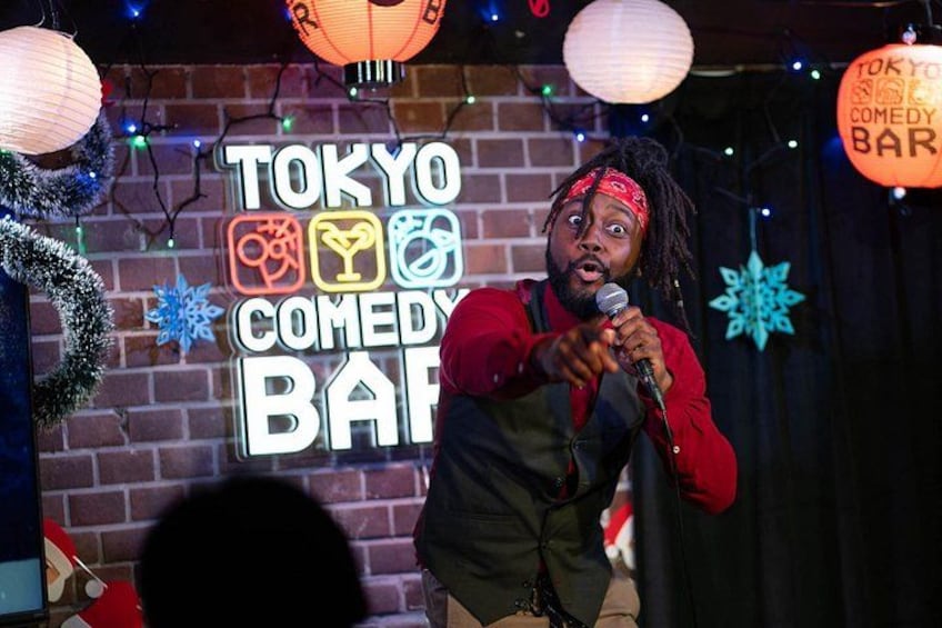 A person performing on stage with a microphone, a brick wall, and a neon sign that reads 'TOKYO COMEDY BAR'. 2