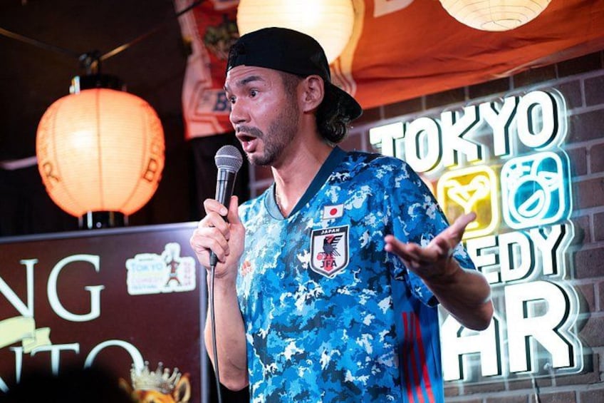 A person in a blue patterned shirt holding a microphone, standing in front of a sign with 'TOKYO' and 'TEDDY' visible. 5