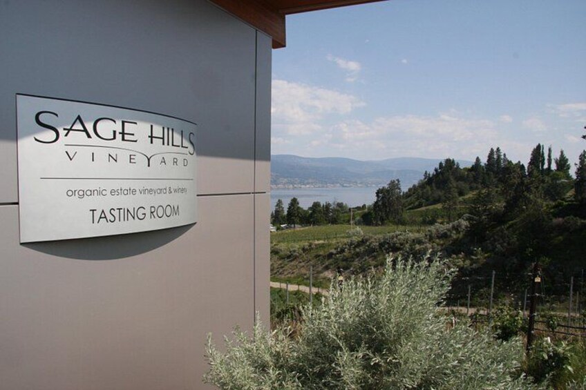 A sign for Sage Hills Vineyard's tasting room with a scenic view of hills and a body of water in the background. 1