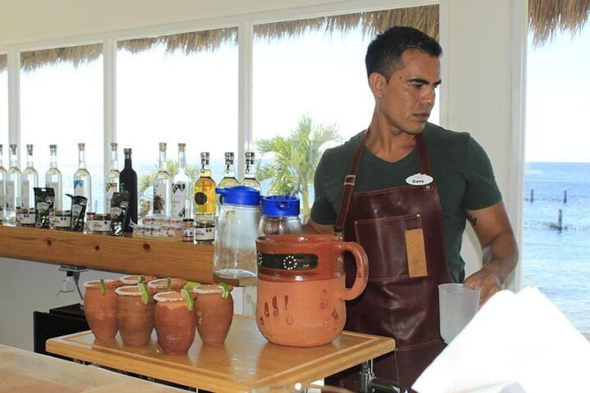 A person in a green shirt and brown apron standing at a bar with various bottles and clay pots. 2