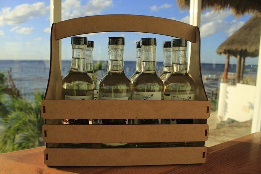 A wooden crate with four bottles of liquor on a wooden table with a view of the ocean and a thatched roof structure in the background. 3