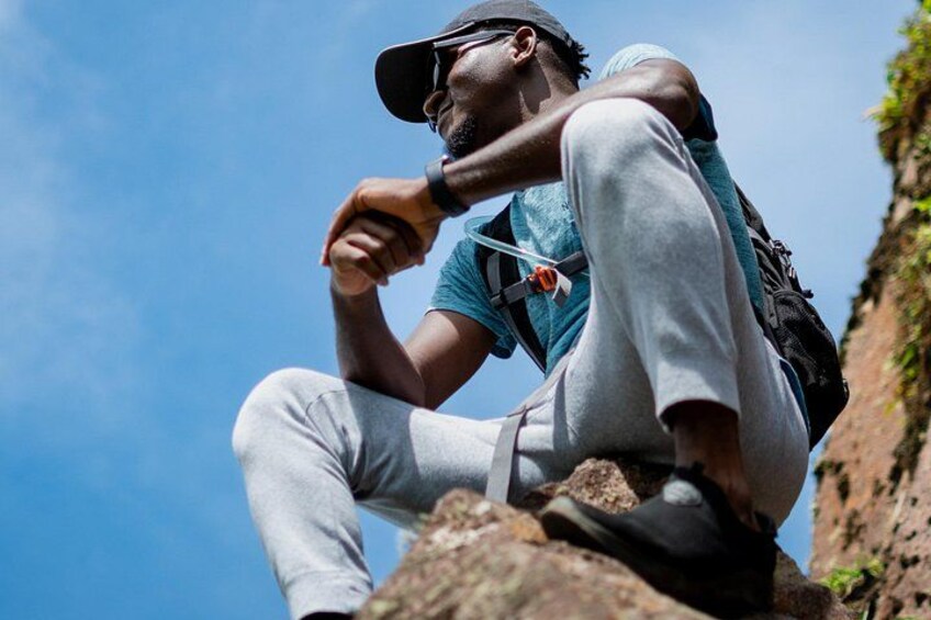 A person sitting on a rock with a backpack, wearing a cap and a watch, against a clear sky. 2