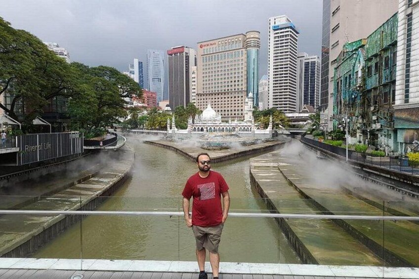 A man standing in front of a water feature with steam rising, surrounded by modern buildings and a traditional temple in the background. 4