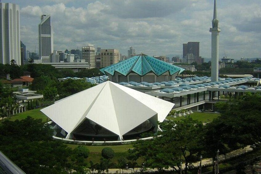 A modern white building with a unique triangular roof, surrounded by lush greenery and a city skyline in the background. 3