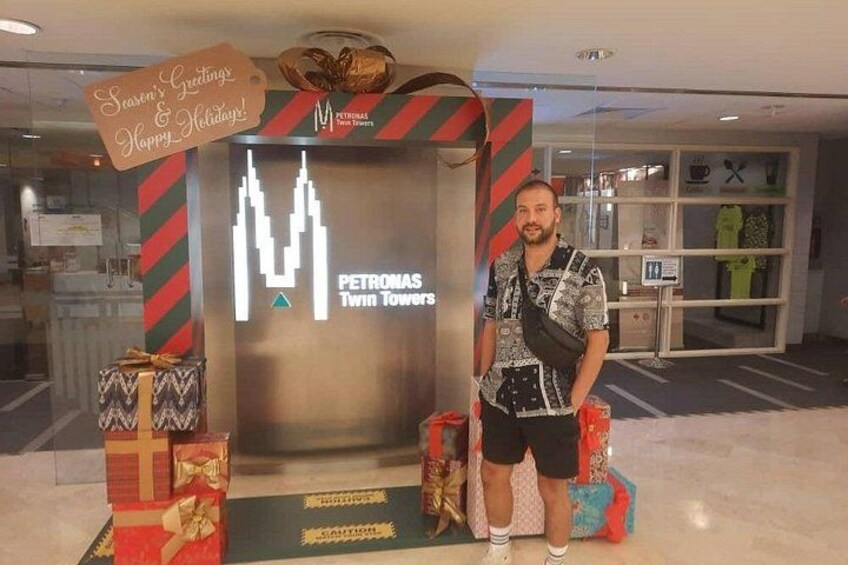 A man standing in front of a PETRONAS Twin Towers display with gift boxes and a holiday greeting sign. 5