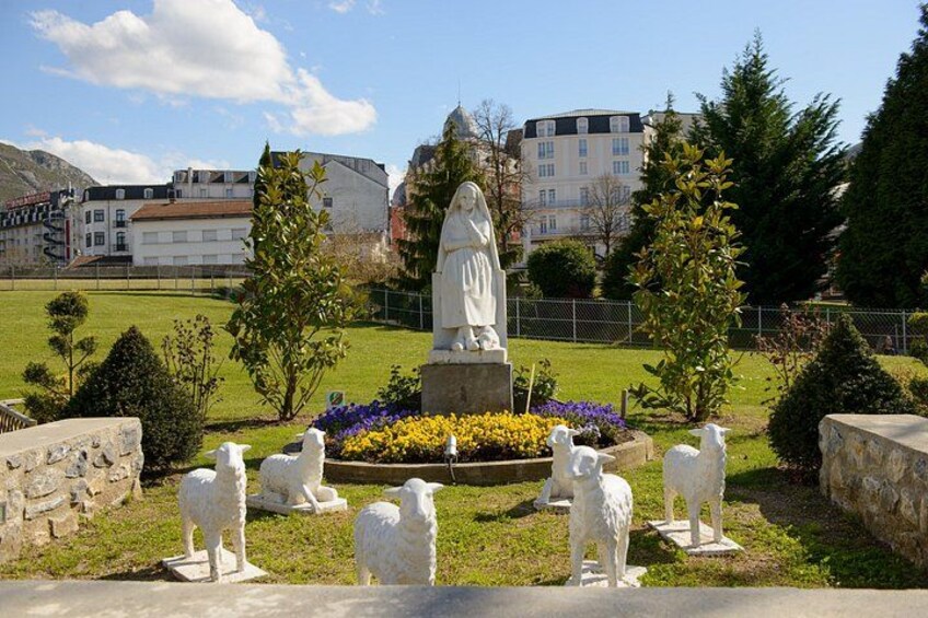 A statue of a woman, sheep sculptures, and a flower bed in a park with buildings in the background. 3