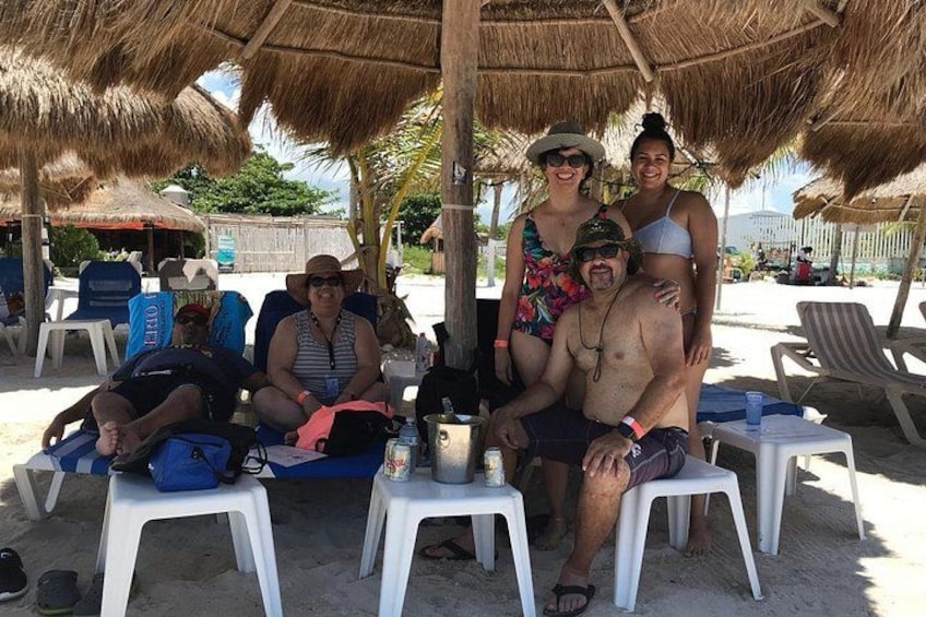 Une scène de plage où l’on voit des personnes assises sur des chaises sous un toit de chaume, avec un palmier et un ciel dégagé. 3