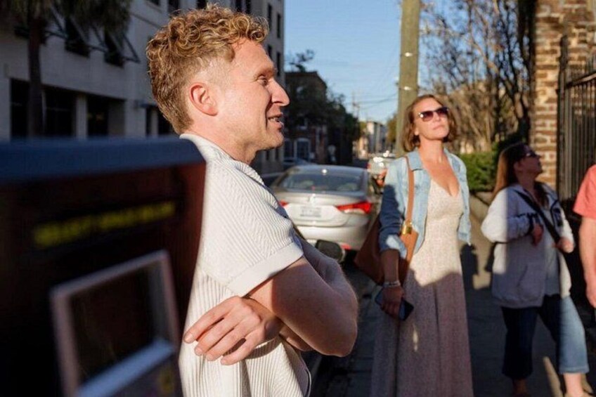 A man standing with his arms crossed, a woman walking in the background, and a car parked on the street. 3