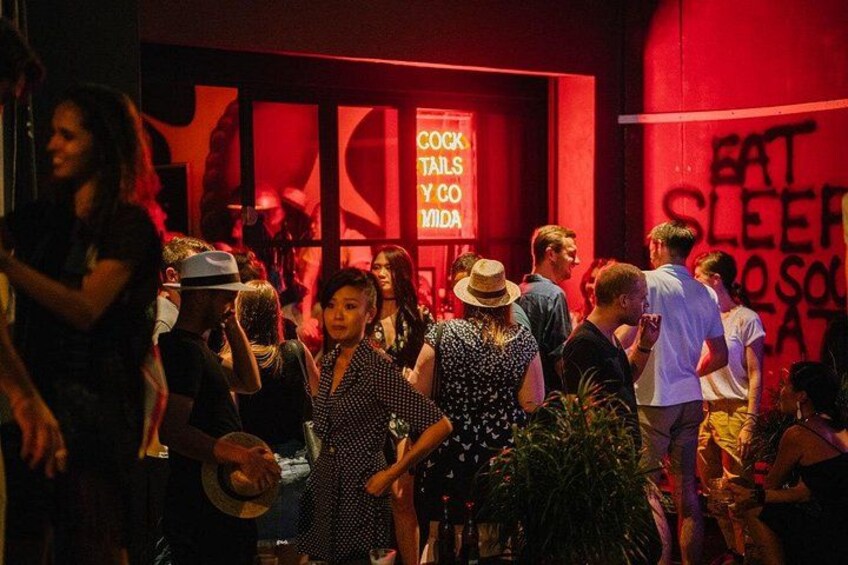 Un evento affollato al chiuso con persone che chiacchierano, una parete rossa con un cartello che recita 'COCKTAILS & COCOA LOVER' e un poster con la scritta 'EAT SLEEP & SOLACE'. 3