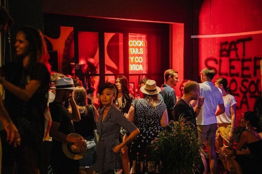 Un événement en intérieur très fréquenté, où les gens se côtoient, un mur rouge orné d’une enseigne sur laquelle on peut lire « COCKTAILS & COCOA LOVER », et une affiche avec le texte « EAT SLEEP & SOLACE ». 3