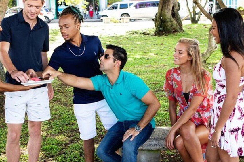 A group of people outdoors, some standing and some sitting, with a man serving food to others. 1
