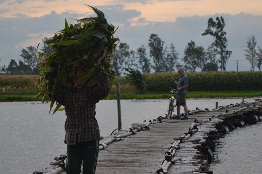 A person carrying a large bundle of plants on a wooden bridge over water, with another person standing nearby. 2