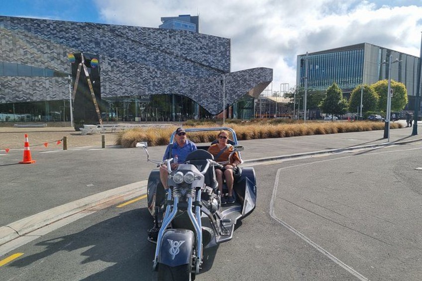 A person riding a motorcycle with a sidecar, another person sitting in the sidecar, and a building with a unique architectural design in the background. 2