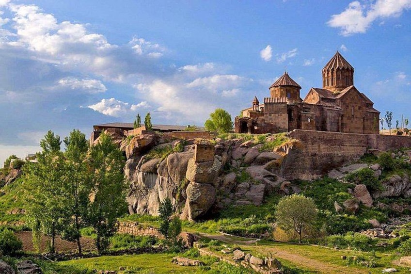 A historic stone building with two domes, situated on a rocky hill with greenery around. 3