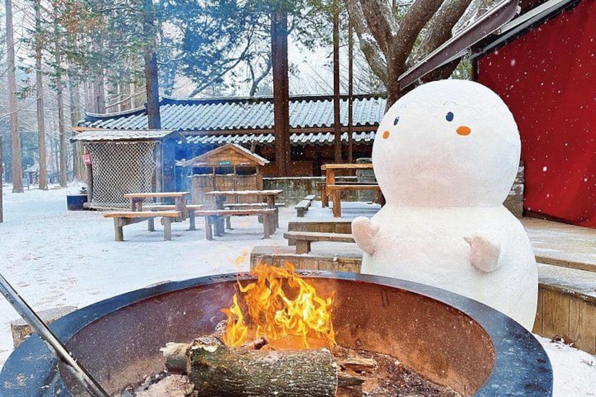 A snowman next to a fire pit in a snowy outdoor setting with traditional wooden structures in the background. 4