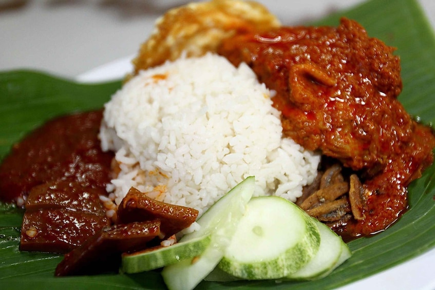 A plate of food with white rice, a red curry, fried chicken, and cucumber slices on a banana leaf. 1