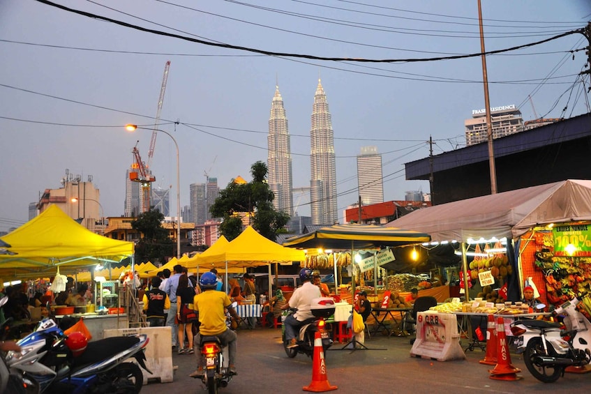A bustling street market with yellow tents and food stalls, with the Petronas Towers in the background. 3