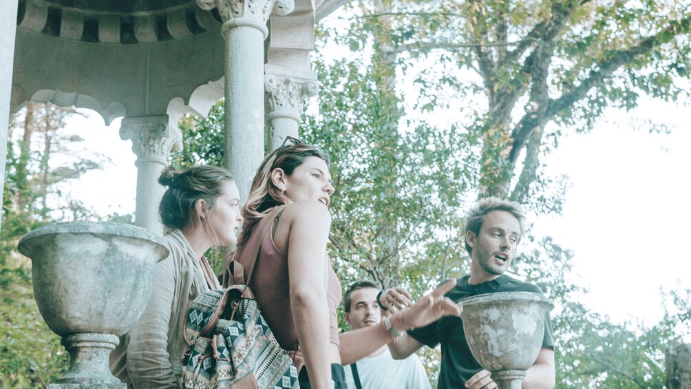 A group of people in an outdoor setting with classical architecture, including columns and urns. 2