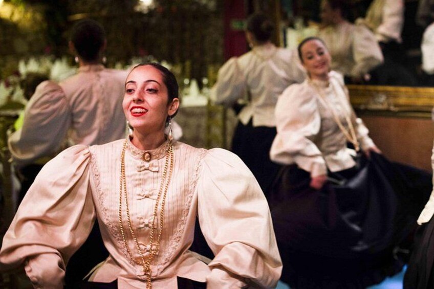 A person in a historical costume with puffed sleeves and a high collar, seated in a room with other individuals in period attire. 4