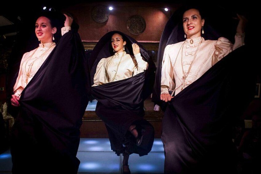 Three individuals in a dimly lit room, wearing long-sleeved, light-colored tops and dark skirts. 2